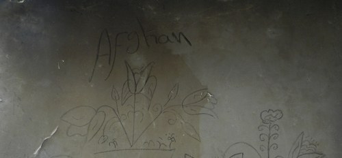 Simple grafiti in black on a dark wall, reads "Afghan" above a simple floral & leaf motif