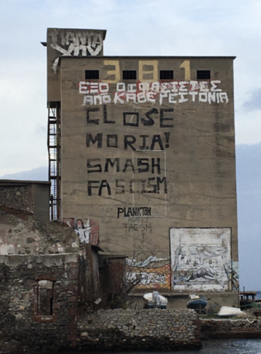 The concrete wall of an  building several stories tall has grafitti in Greek and English. The English reads in black, bold letters: Close Moria! Smash Facism! The building is right on the water, and several small boats are on land