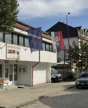 A white administrative builting with a Serbian flag over the entrance and a sign that says "registration" in English, and has Arabic text below,