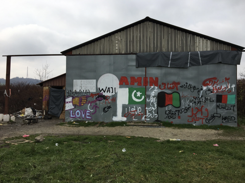 A barn like building is covered with grafitti in many languages including images of the flags of Afghanistan and Pakistan, and a hand pointing with one finger to the right