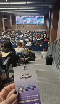 Vasiliki's conference badge is visible in the foreground; the background shows, from the back, an auditorium full of people